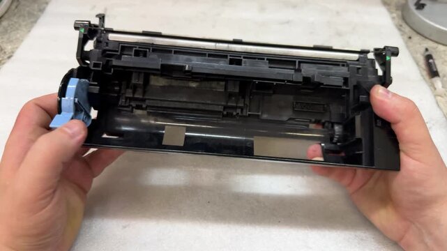 Two hands hold a black developer unit of a printer cartridge with a blue gear. Printer repair, spare parts, device service, office equipment