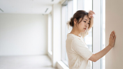 Naklejka premium A young asian woman standing by a window in a bright, minimalist room, looking tired or stressed with her hand on her forehead