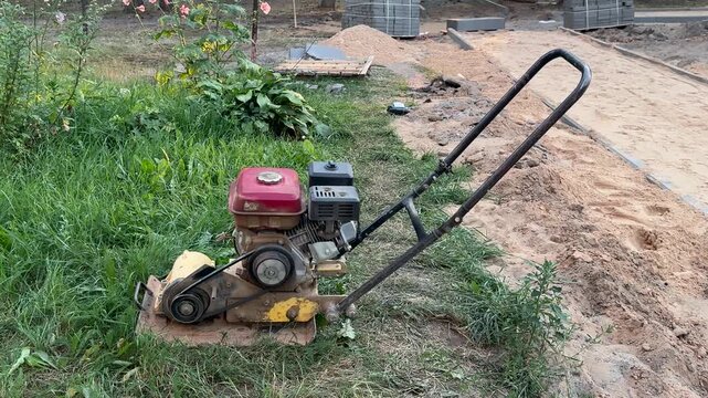 A gasoline plate compactor stands on the grass next to sand at a paving site. Road construction, paving work, site equipment, ground compaction