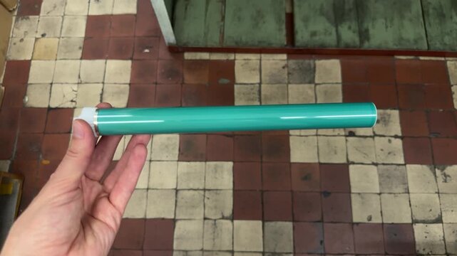 A hand holds a green cylindrical photoconductor drum for a printer cartridge. Printer repair, spare parts, device service, office equipment