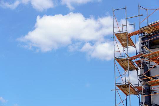 Construction scaffolding against a clear blue sky with fluffy white clouds. Metal framework with wooden platforms on the side of a modern building facade.