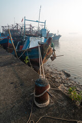 Weathered blue wooden fishing boats tied to a rusted mooring bollard with thick ropes at a calm harbor during a hazy morning sunrise © Zaenal