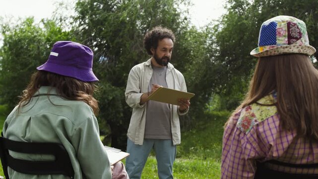 Middle aged man teaching outdoor drawing class to young adult women. Footage is ideal for promoting creative workshops, art education programs, or community events