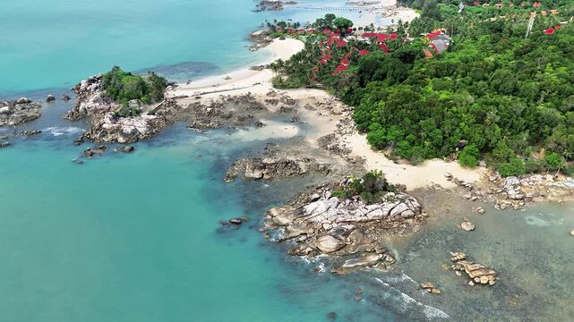 Aerial view of tropical paradise beach with crystal turquoise ocean, white sand shoreline, rocky coast formations, and lush green forest creating peaceful island scenery