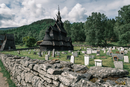 View of Borgund Stave Church with dark timber architecture, surrounded by a historic cemetery with stone grave markers, a dry stone wall, and green trees in Borgund, Vestland, Norway.