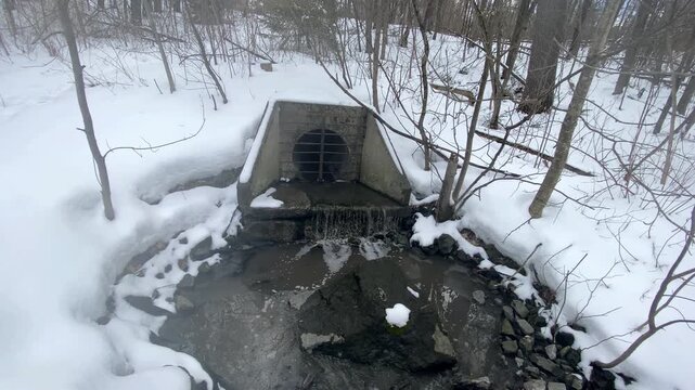Concrete culvert headwall discharging stormwater runoff into a rocky creek bed surrounded by snow-covered winter forest and bare deciduous trees.