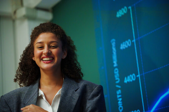 Smiling businesswoman speaker giving a professional seminar presentation beside a digital financial chart, representing leadership, market analysis, confidence, training and business success.
