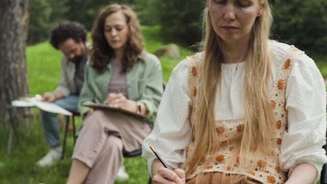 Caucasian young adult woman drawing in sketchbook during outdoor art class, camera tilting up to her face. Video is perfect for promoting creative workshops, art education, and hobbies