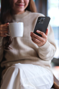Vertical closeup image of a woman holding and using mobile phone while drinking coffee in cafe
