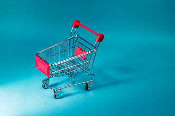 An empty small metal shopping cart with red handles set against a blue gradient background for commerce and sale layouts. High Quality Photo © 7TridentSea