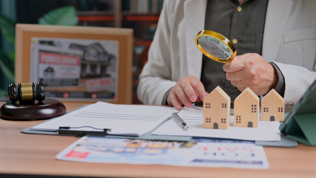 Person inspects wood home models desk docs gavel magnifier real estate agency law judge analyzing property contract details buyer transaction mortgage investment strategy legal risk assessment tax.