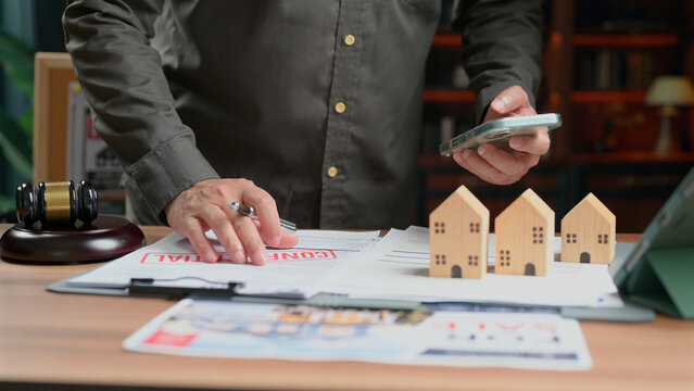 Person checking mobile device inspecting real estate contract documents wooden house models office desk lawyer verifying property agreement buyer checking mortgage loan terms business paperwork.