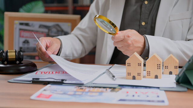 Person carefully examines real estate contract document holding magnifying glass wooden house models gavel office desk legal transaction agreement purchasing housing assets investment planning.