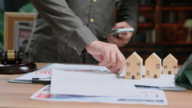Individual reviewing real estate contract holding pen near wooden house models auction gavel smartphone workspace table mortgage agreement property purchase.