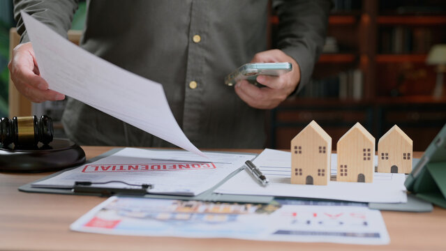 Agent inspects confidential property contract document near miniature wooden house models court gavel desk smartphone holding loan agreement paper legal consulting asset management corporate finance.