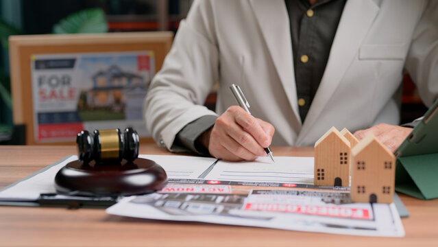 Individual signing legal contract document beside miniature house model judge gavel resting wooden desk office executing property purchase agreement transaction loan approval business commercial.