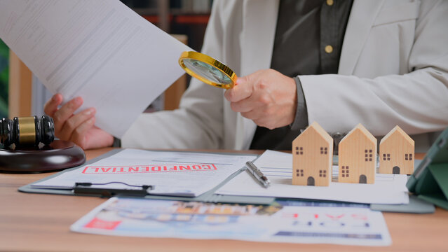 Person wearing suit examines confidential document holding magnifying glass desk featuring wooden house models gavel contract paperwork legal investigation real estate market analysis corporate.
