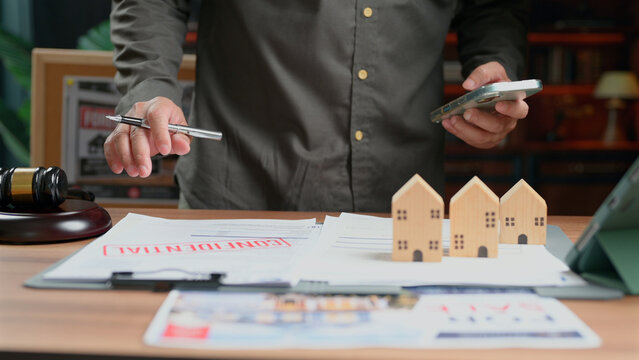 Man reviewing real estate contract documents holding pen smartphone wooden house models desk legal gavel office setting lawyer agent property transaction insurance claim paperwork business.