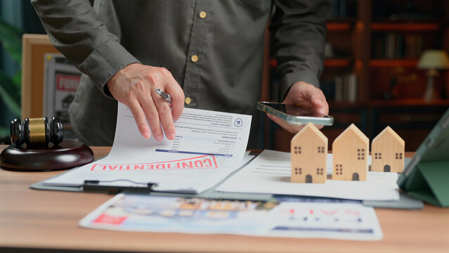 Man checking legal paper in office holding pen phone near house model gavel representing property deal business agreement purchase transaction mortgage loan scheme investment asset fund portfolio.