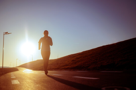 Silhouette Of Female Runner Jogging On Road At Golden Sunset. Athletic young woman running outdoors towards the sun during beautiful evening workout
