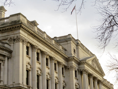 View of the neoclassical facade of HM Treasury with its grand columns and a flag flying against a cloudy sky in London, England, United Kingdom.