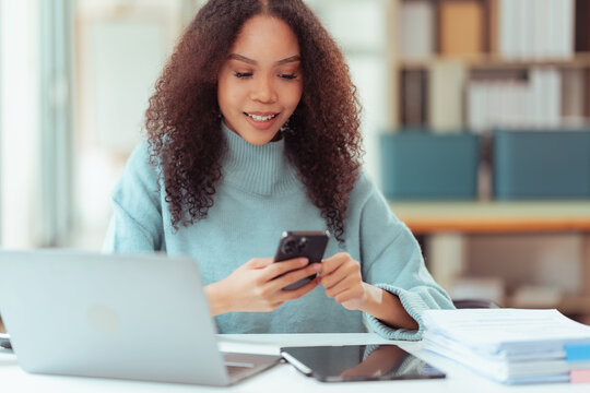Happy Young African American Woman Using Smartphone at Desk with Laptop and Tablet