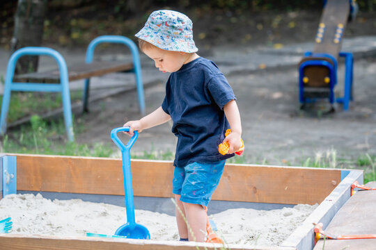 Little boy playing with a large blue shovel in a sandbox outdoors.