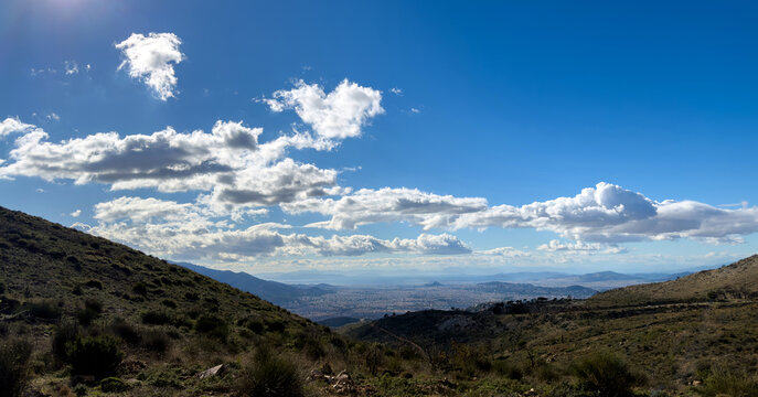 Rocky scrub covered slopes overlooking urban valley under cumulus clouds in Penteli, Attica Greece