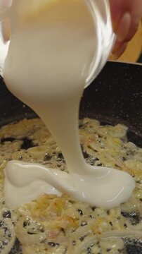 Hand Pours Thick White Sauce Over Sauteed Onions in a Black Frying Pan, Close Up View of Creamy Food Preparation Slow motion Vertical video. Vertical video.