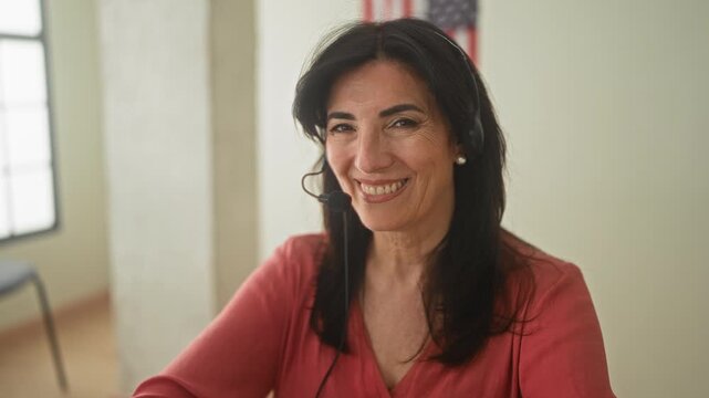 Middleaged hispanic woman wearing a headset smiling beside an american flag inside a building; friendly support.