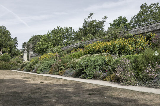 View of a lush, vibrant flower border in a historic garden with a stone wall and gravel path in Oxford, England, United Kingdom.