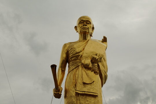 Golden Buddhist monk statue holding fan against cloudy sky