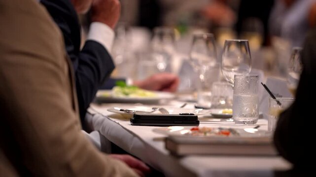 Footage of a formal business dinner with people in suits seated at a table with glassware and white tablecloth, creating an abstract corporate atmosphere.
