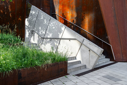 Beside a metal wall marked by rust, concrete stairs rise through dense plants as greenery and landscaping frame the path within layered urban architecture.