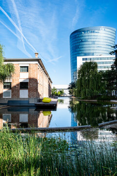 Along a canal, buildings meet blue sky as vegetation borders the water and reflection extends modern urban architecture beside a sunlit facade.