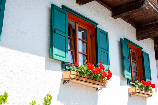 Windows with Green Shutters and Red Geraniums on White Facade of Alpine House at Sunny Summer Day.