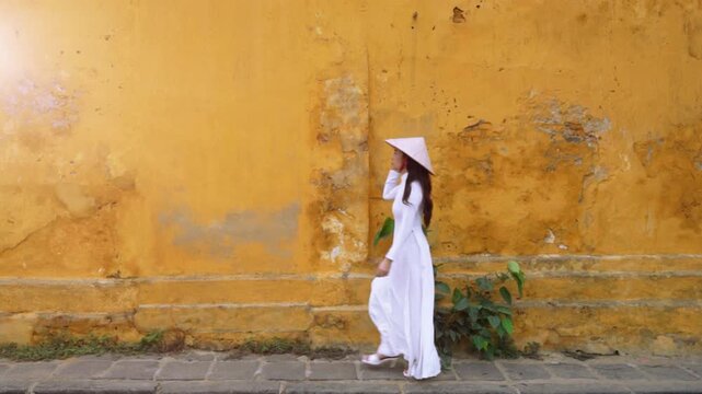Asian woman wearing traditional Vietnamese culture walks beside an old wall in Hoi An, Vietnam.