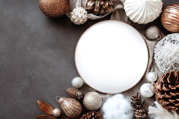 Obraz premium Round white plate surrounded by assorted decorative Christmas ornaments, pine cones, and natural elements on a dark textured surface for holiday-themed displays