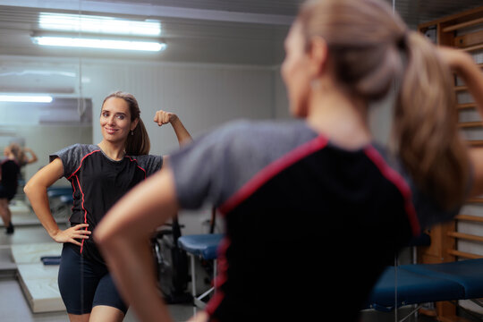 Strong Female Patient Flexing Muscle In Mirror At Rehabilitation Center. Smiling young woman looking at reflection showing biceps during physical therapy recovery