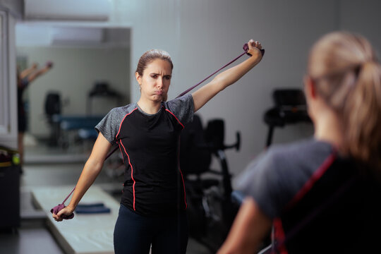 Struggling Female Patient Using Resistance Band At Rehabilitation Center Mirror. Young woman making funny face while performing physical therapy shoulder exercise outdoors