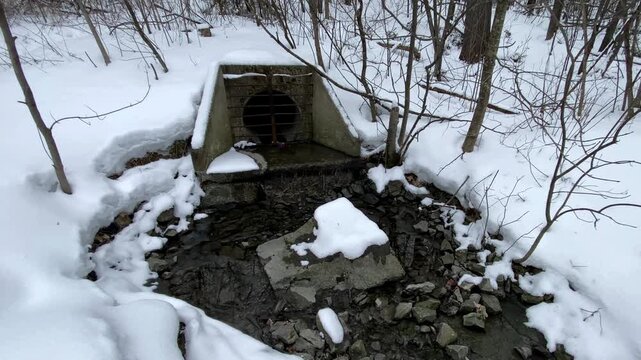 Concrete culvert headwall discharging stormwater runoff into a rocky creek bed surrounded by snow-covered winter forest and bare deciduous trees.