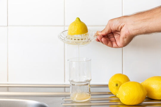Jus de citron frais press&eacute; avec un presse agrume en verre, une main qui verse le jus dans un grand verre transparent pos&eacute; sur un &eacute;vier en inox et trois citrons pos&eacute;s &agrave; cot&eacute;, carrelage blanc en fond