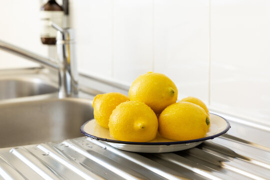 Des citrons jaunes sur une assiette &eacute;maill&eacute;e blanche avec un liser&eacute; bleu pos&eacute;e sur un &eacute;vier en inox dans une cuisine, carrelage blanc en arri&egrave;re-plan, vue en perspective