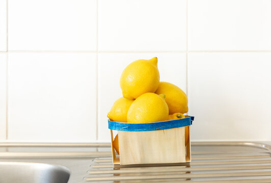 Des citrons jaunes empil&eacute;s dans une barquette en bois avec un liser&eacute; bleu pos&eacute;e sur un &eacute;vier en inox dans une cuisine, carrelage blanc en arri&egrave;re-plan