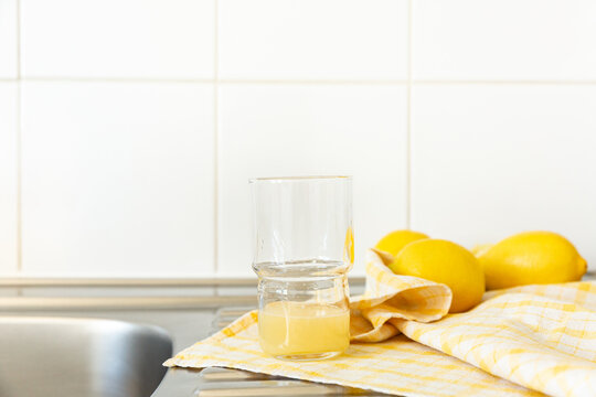 Jus de citron frais press&eacute; dans un grand verre transparent pos&eacute; sur un torchon &agrave; carreaux jaunes et blancs, trois citrons entiers en arri&egrave;re-plan, &eacute;vier de cuisine en inox et carrelage blanc