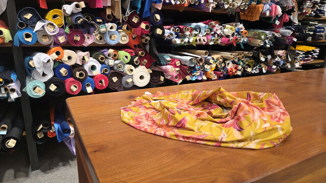 Draped yellow floral fabric on wooden counter with organized fabric bolt shelves in background for shop display and banner