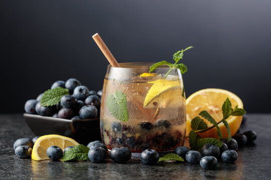 Iced blueberry drink with mint, lemon, ice, and fresh berries on a black stone table.