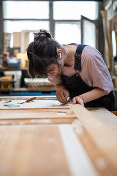 Craftswoman engaged in working process in carpentry, master sanding wooden door with sandpaper, female restorer carefully polishing and smoothing surface, removing roughness and defects of putty.