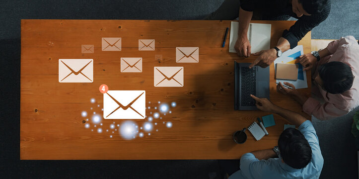 A top view of a collaborative office setting featuring professionals engaged in discussion around a wooden table, complemented by visible email notifications and icons. Trope