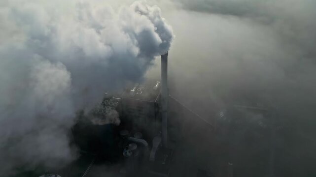 Aerial view of an industrial plant with a tall chimney releasing thick smoke into the surrounding fog and low clouds in Latvia.